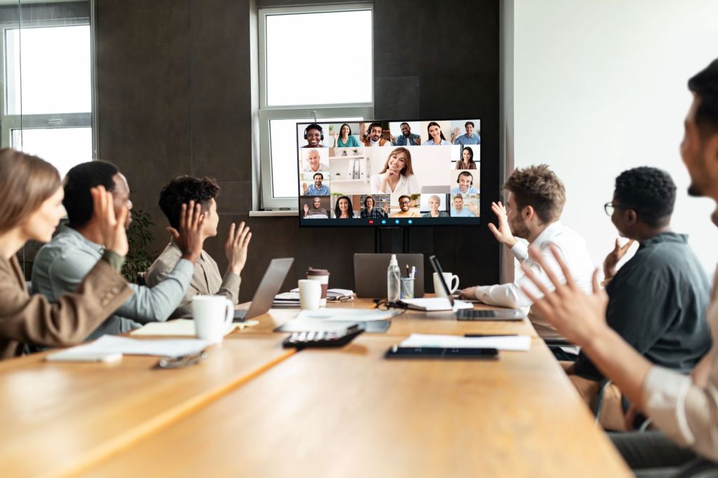 Distant Virtual Meeting Concept. Diverse business people making online video call with colleagues, sitting at desk in boardroom, waving hands at tv screen at office, talking on web with remote workers